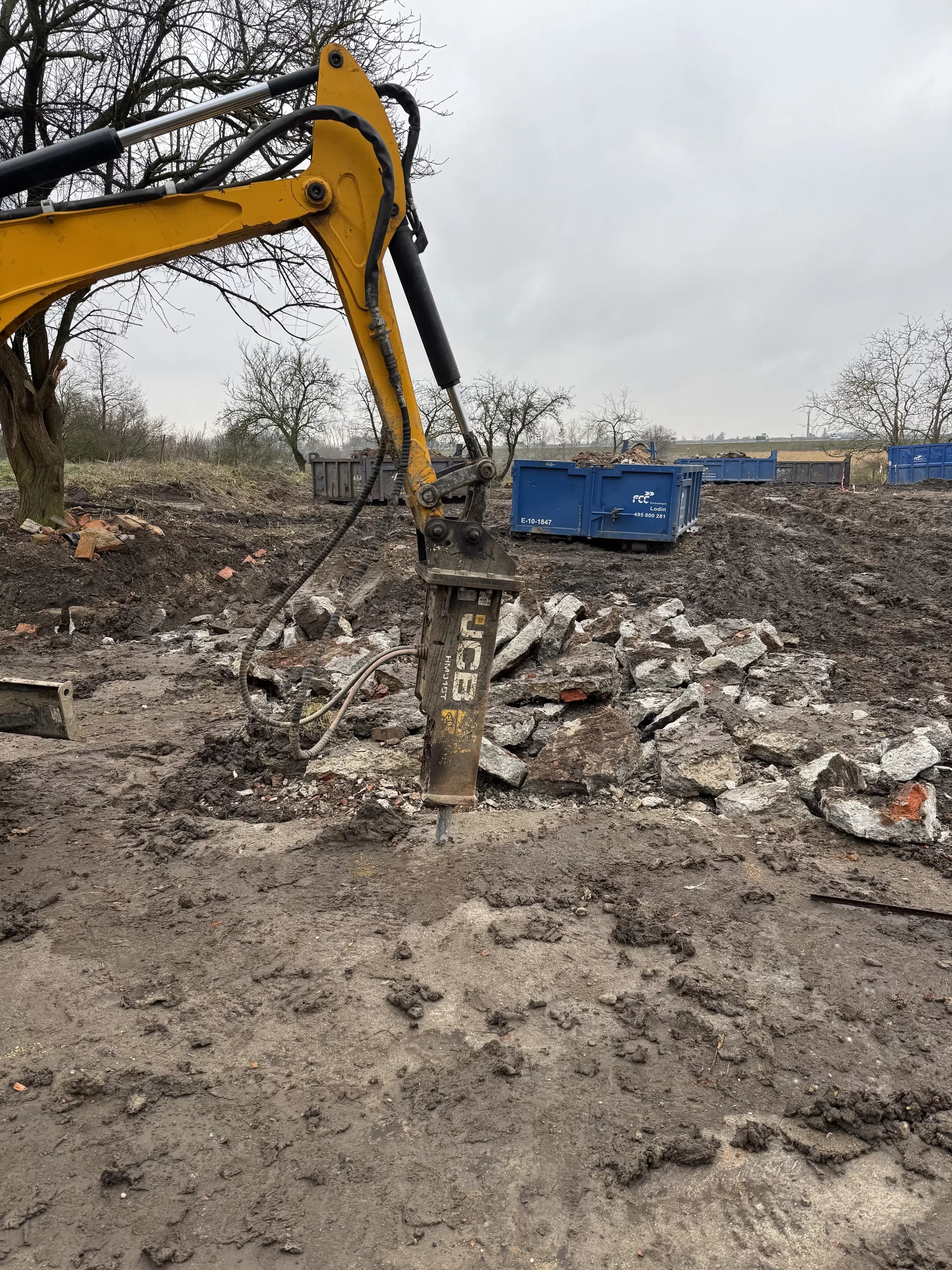 Bourací kladivo na bagru rozbíjí betonové základy, v pozadí kontejnery